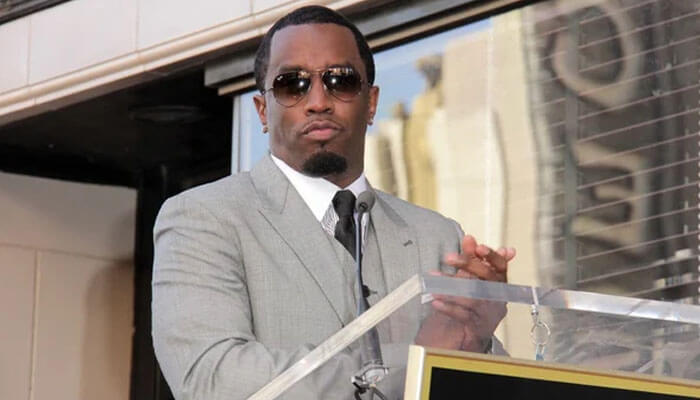 Man in grey suit and sunglasses at podium, clapping—used to illustrate the phrase nice try Diddy meaning in pop culture context.