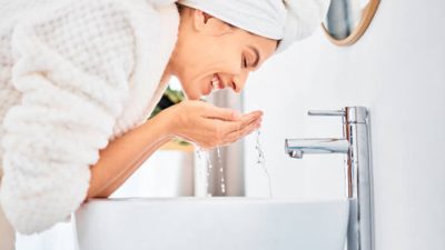 Person washing face in a modern bathroom, smiling with water flowing from their hands, showcasing a refreshing skincare routine