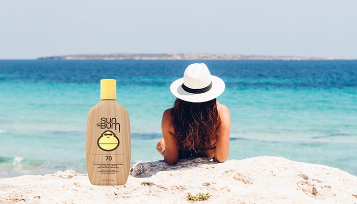 Sun Bum SPF 70 sunscreen bottle placed next to a woman relaxing on the beach, with turquoise waters in the background. Is Sun Bum sunscreen safe for your skin and the environment?