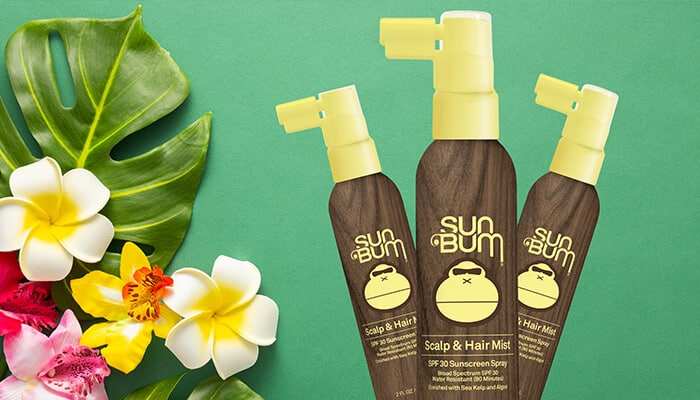 Three Sun Bum Scalp & Hair Mist bottles with SPF 30, surrounded by tropical flowers. Is Sun Bum sunscreen safe for your scalp and hair protection?