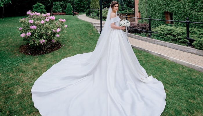 Bride in a classic off-shoulder satin A Line Wedding Dress with a long flowing train in a garden setting.