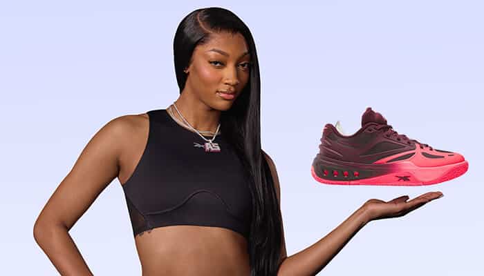 Angel Reese posing in a black athletic top while holding a bright pink Reebok sneaker from the Angel Reese Shoes collection in her hand.
