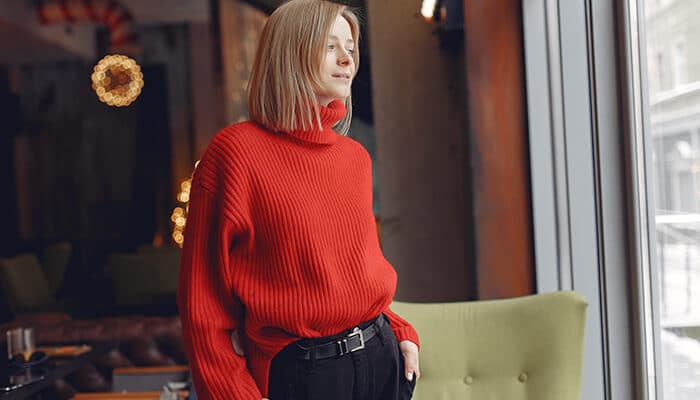 A woman wearing a vibrant red cashmere sweater, styled with black pants, standing by the window, representing stylish and comfortable best cashmere sweaters.