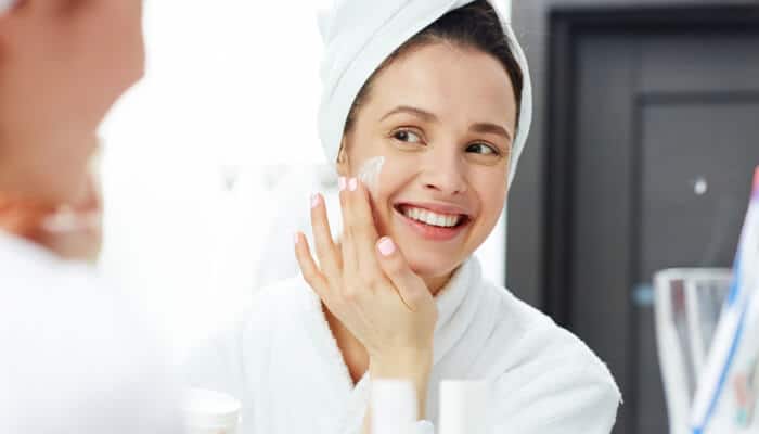 Moisturizer - Fashion in beauty - Fashion in Beauty How to get clear skin: Woman applying moisturizer to her face, smiling in front of a mirror, following a skincare routine for smooth and healthy skin.