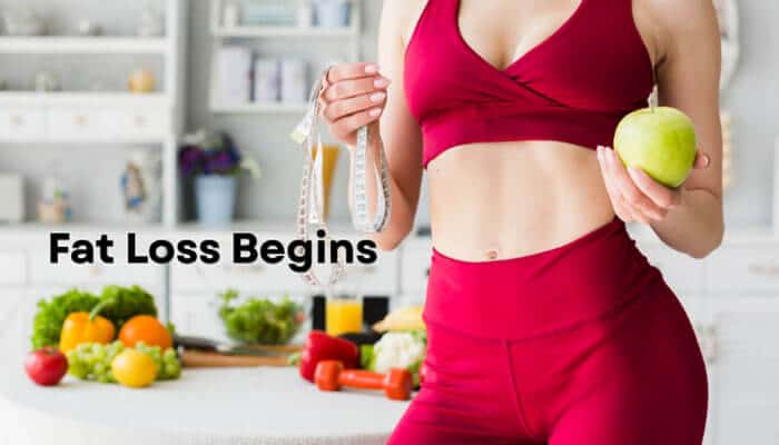 Fat Loss Begins - Fashion In Beauty - Fashion in Beauty Fit woman in red activewear holding a green apple and measuring tape with fresh fruits and dumbbells on kitchen counter, illustrating Tirzepatide Weight Loss and fat loss progress.
