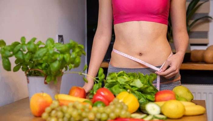Maintain a Comfortable Weight - Fashion In Beauty - Fashion in Beauty Woman measuring her waist beside fresh fruits and vegetables, highlighting healthy weight management as a Home Remedy for Acid Reflux at Night.